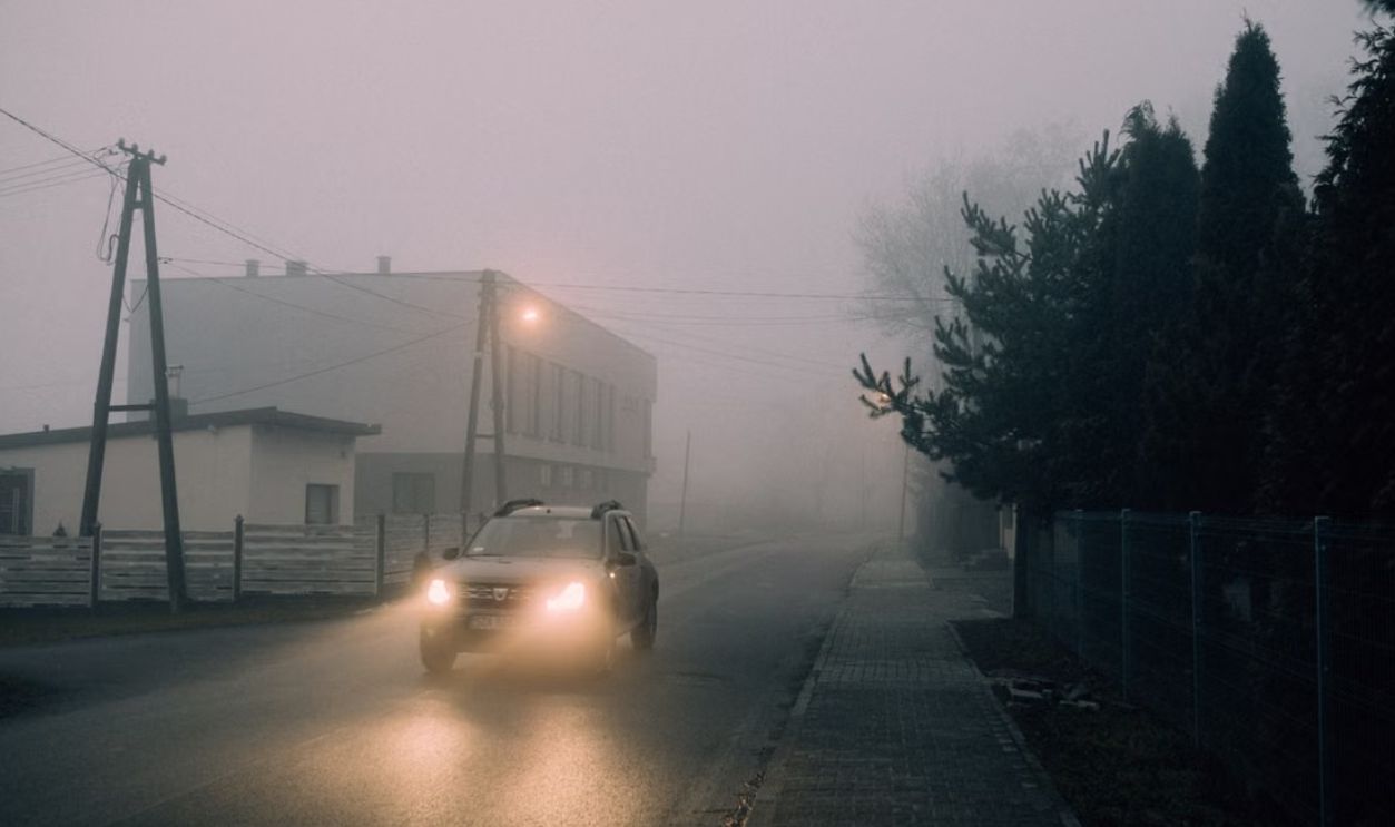 Moody scene of a car driving through a foggy street at night with glowing headlights