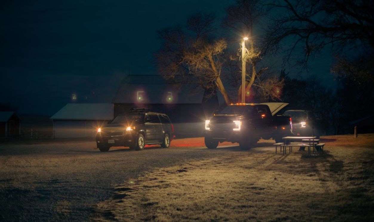 Vehicles with headlights on parked near a barn, illuminated by light post at night