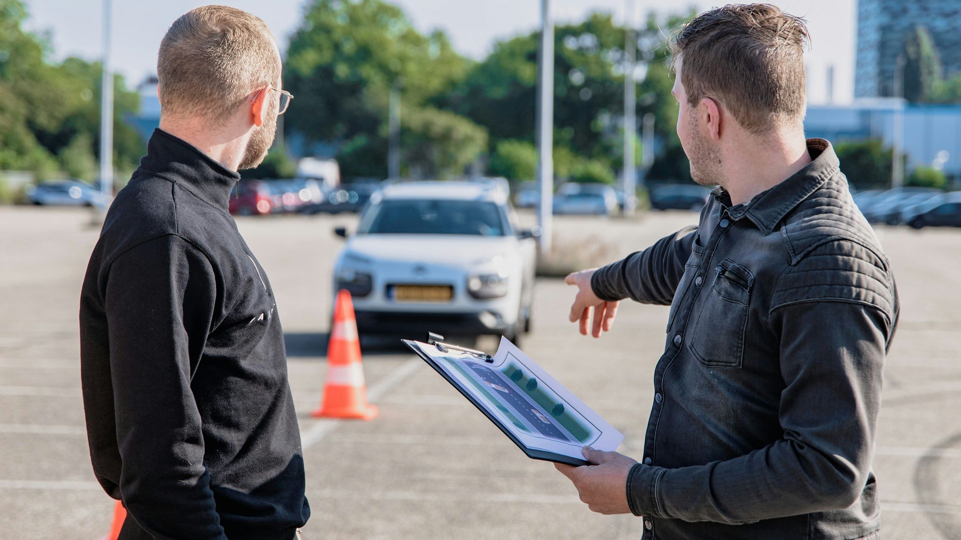 A man pointing toward a car while holding a clipboard for a driving test