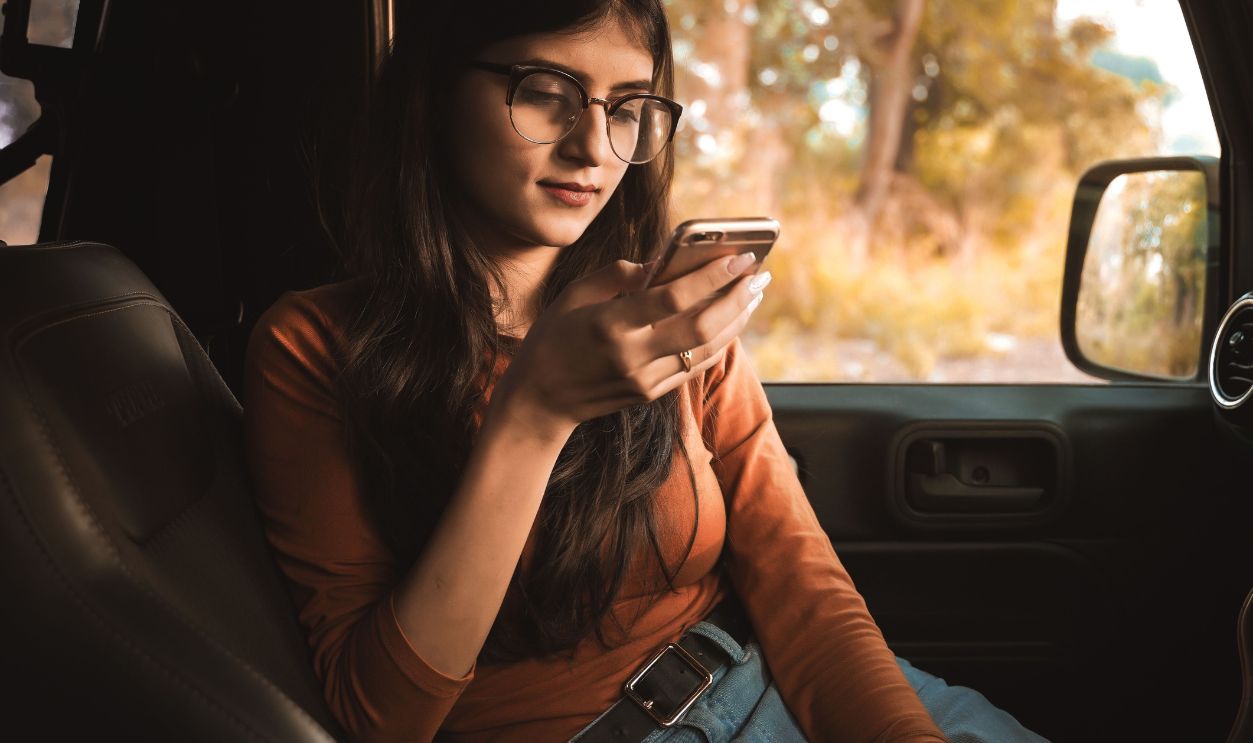 A young woman wearing glasses and casual clothing relaxes in a car, using her phone on a sunny day