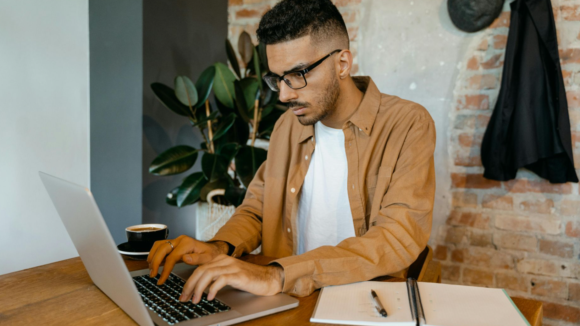 Man in casual office setting typing on laptop with coffee and notebook on desk.