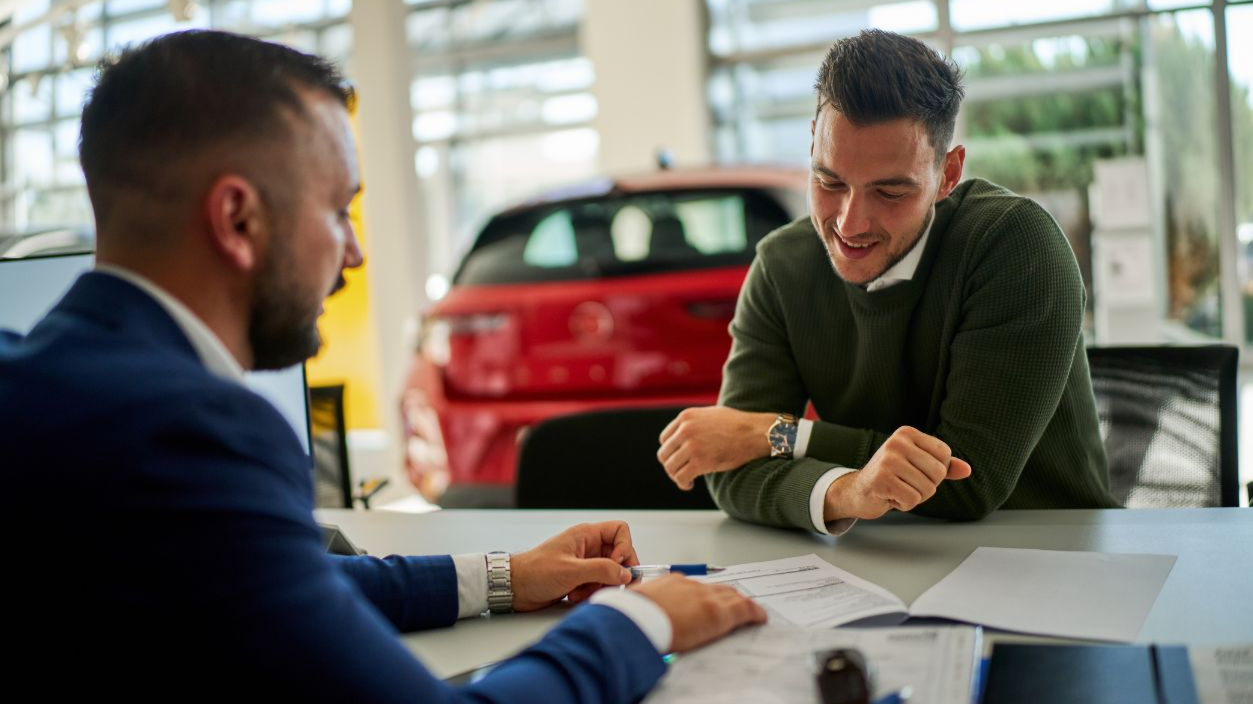 Shutterstock-2582442077, Car salesman and a customer engage in a discussion at a dealership table. Documents and a car key are on the table, with a vehicle visible in the background through large windows