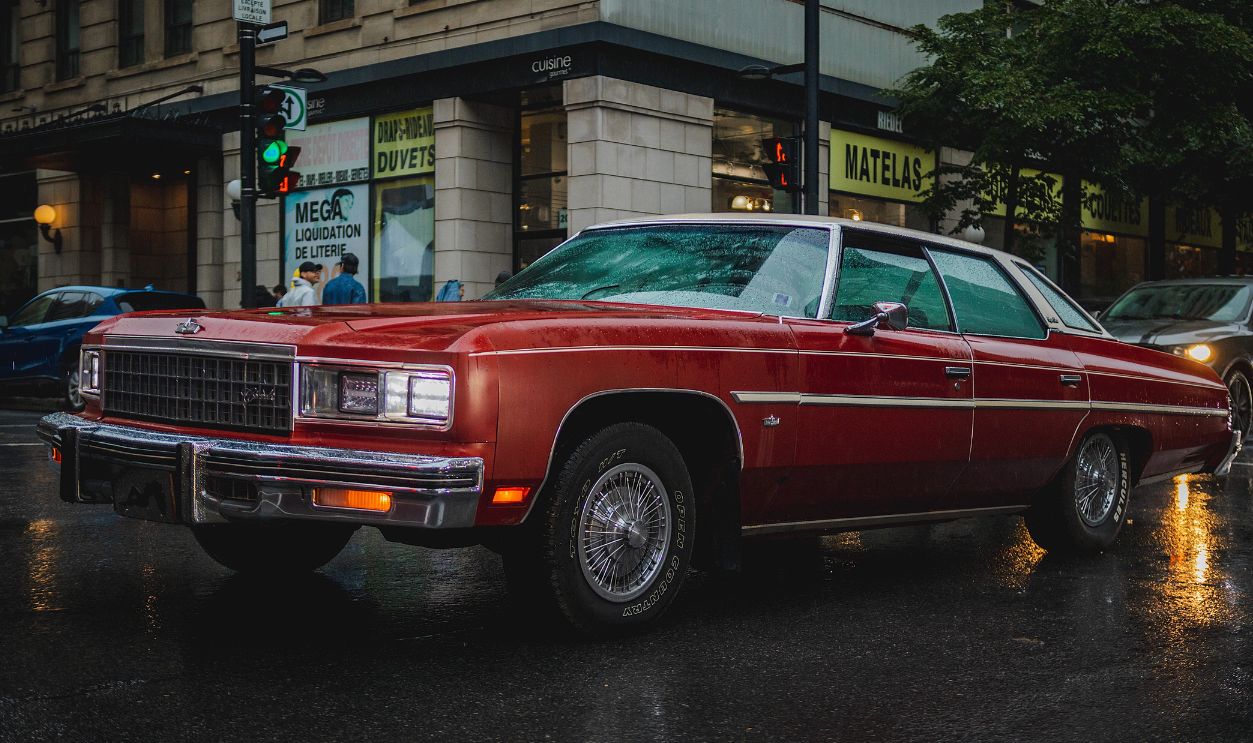 A red vintage car parked on a rainy city street, reflecting urban nostalgia