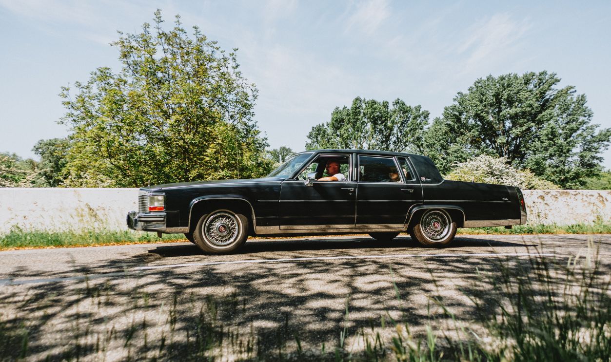 Vintage black sedan cruising on a scenic road on a sunny day, surrounded by lush greenery