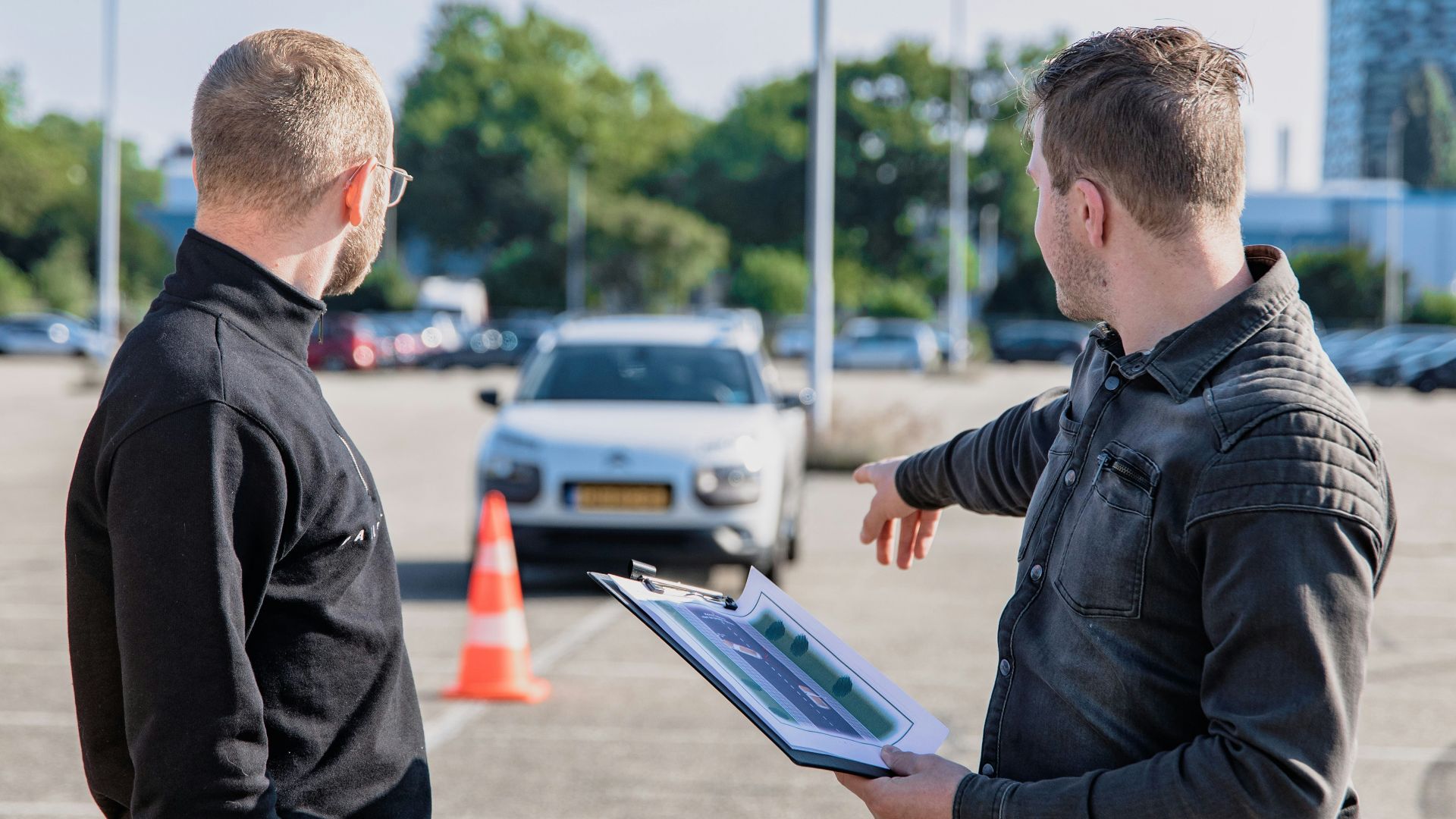 A man pointing toward a car while holding a clipboard for a driving test