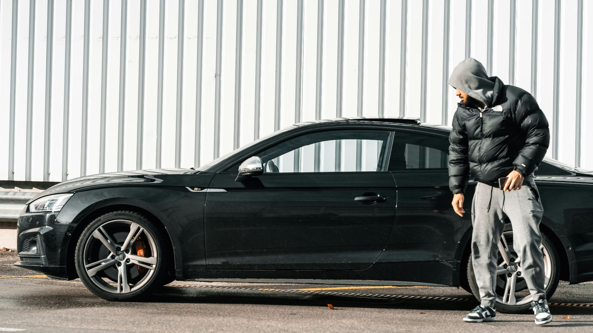 a man in a black jacket and hoodie leaning against a black car