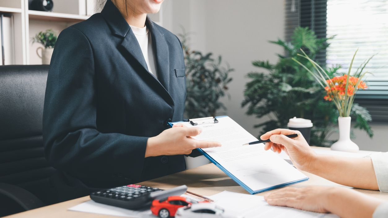 Shutterstock-2750734997, Smiling agent explaining auto loan contract to client at office desk with calculator and model cars, representing vehicle purchase consultation, payment planning, insurance options and trust.
