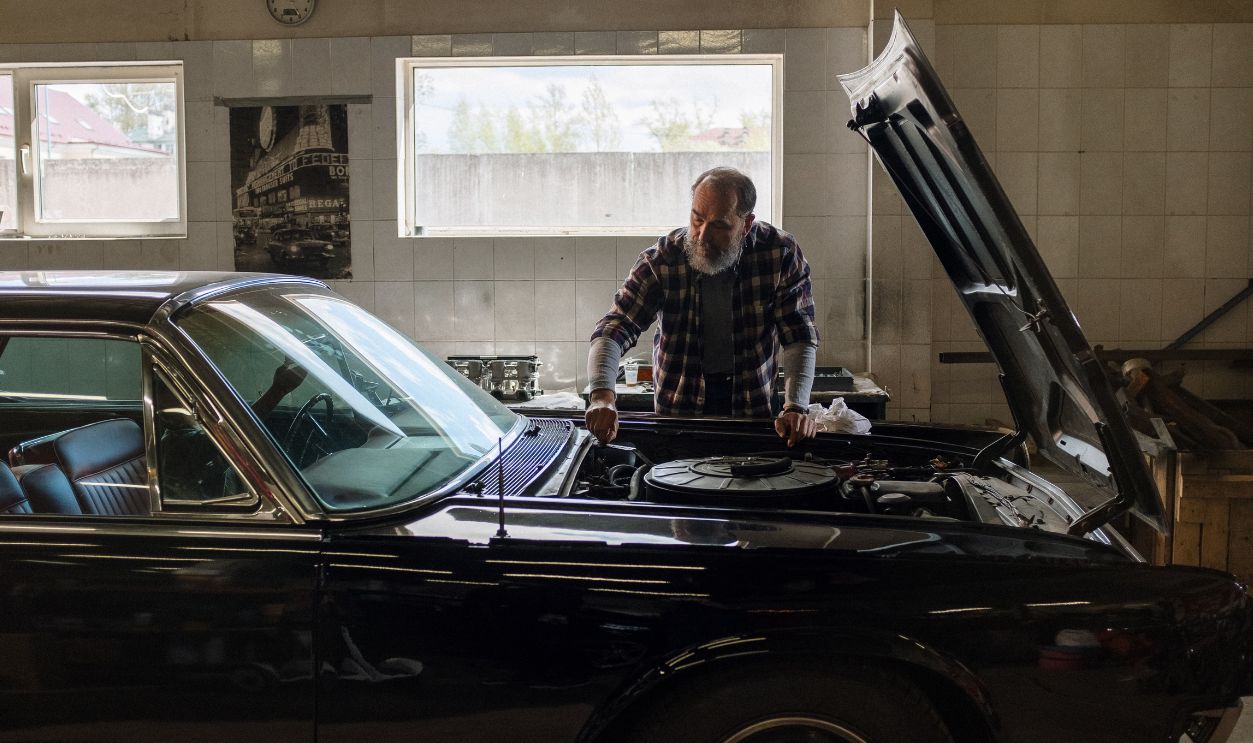 A bearded mechanic examines a classic car engine in a well-lit auto repair shop