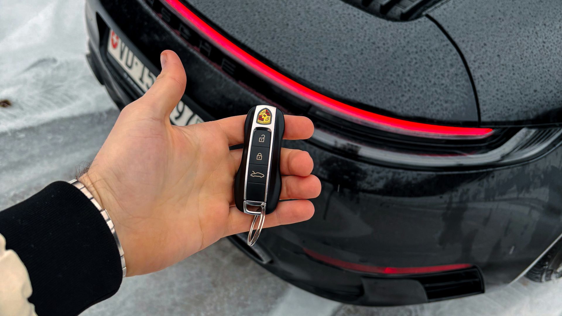 Hand holding car key in front of black porsche