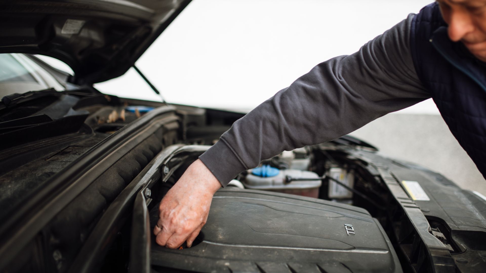 A person is leaning over an open car hood in a garage. They focus on the engine, checking parts and doing maintenance. Tools and equipment are visible around them.