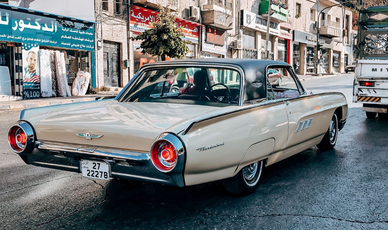 A vintage Thunderbird car drives down a vibrant urban street lined with shops.