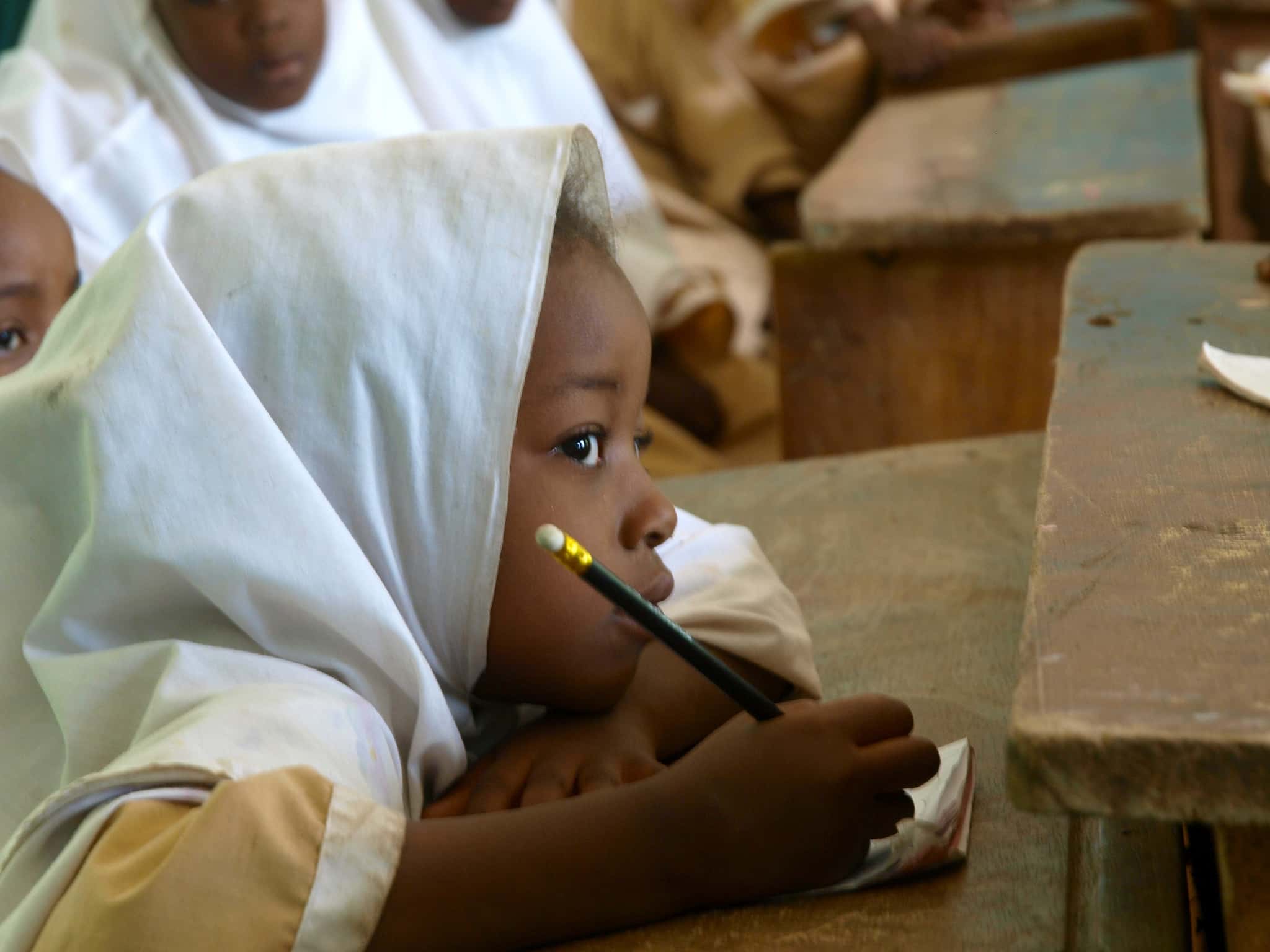 Child wearing white clothes holding pencil sitting on a school desk