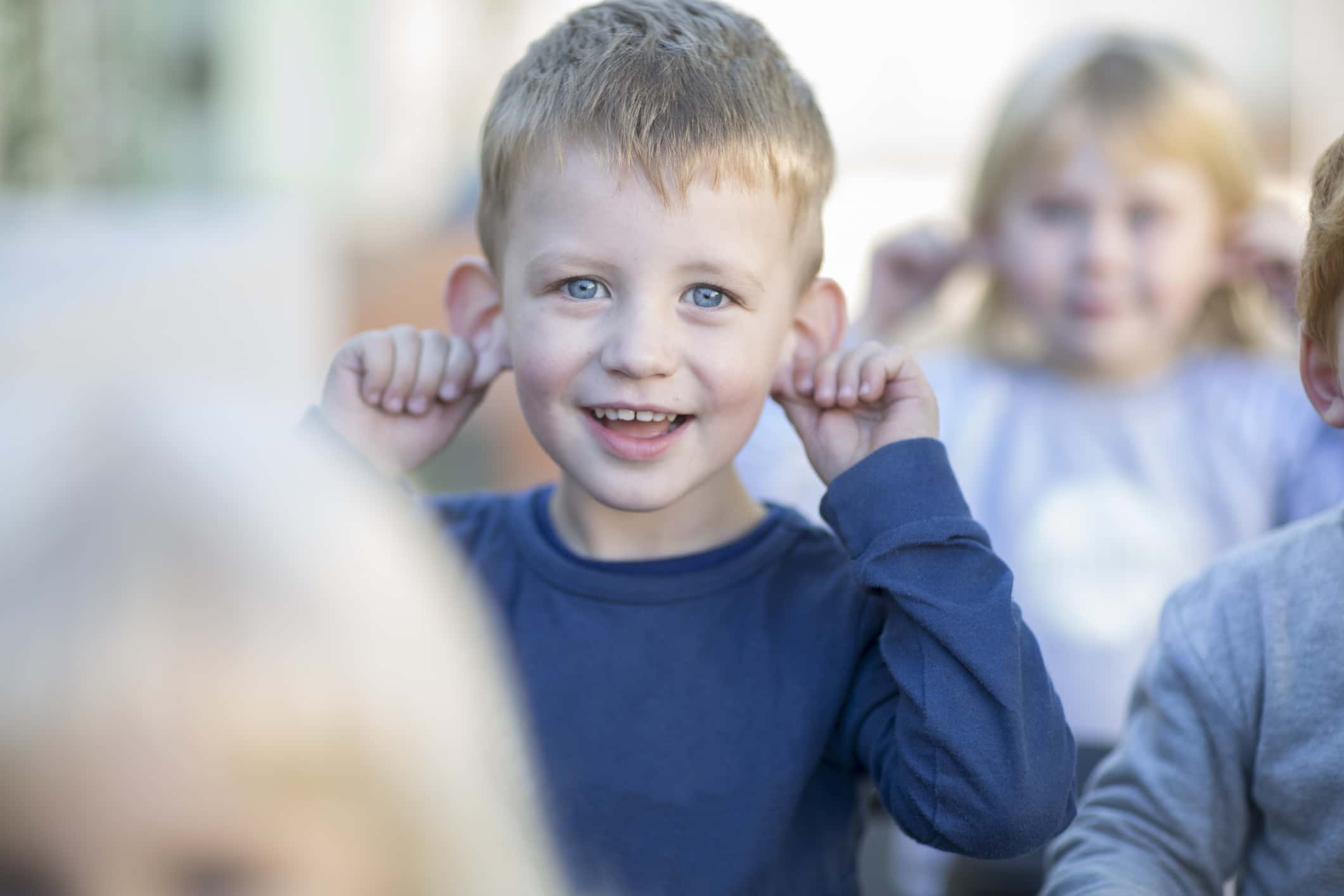 Portrait of young boy wearing a blue shirt, outdoors, holding ears, smiling