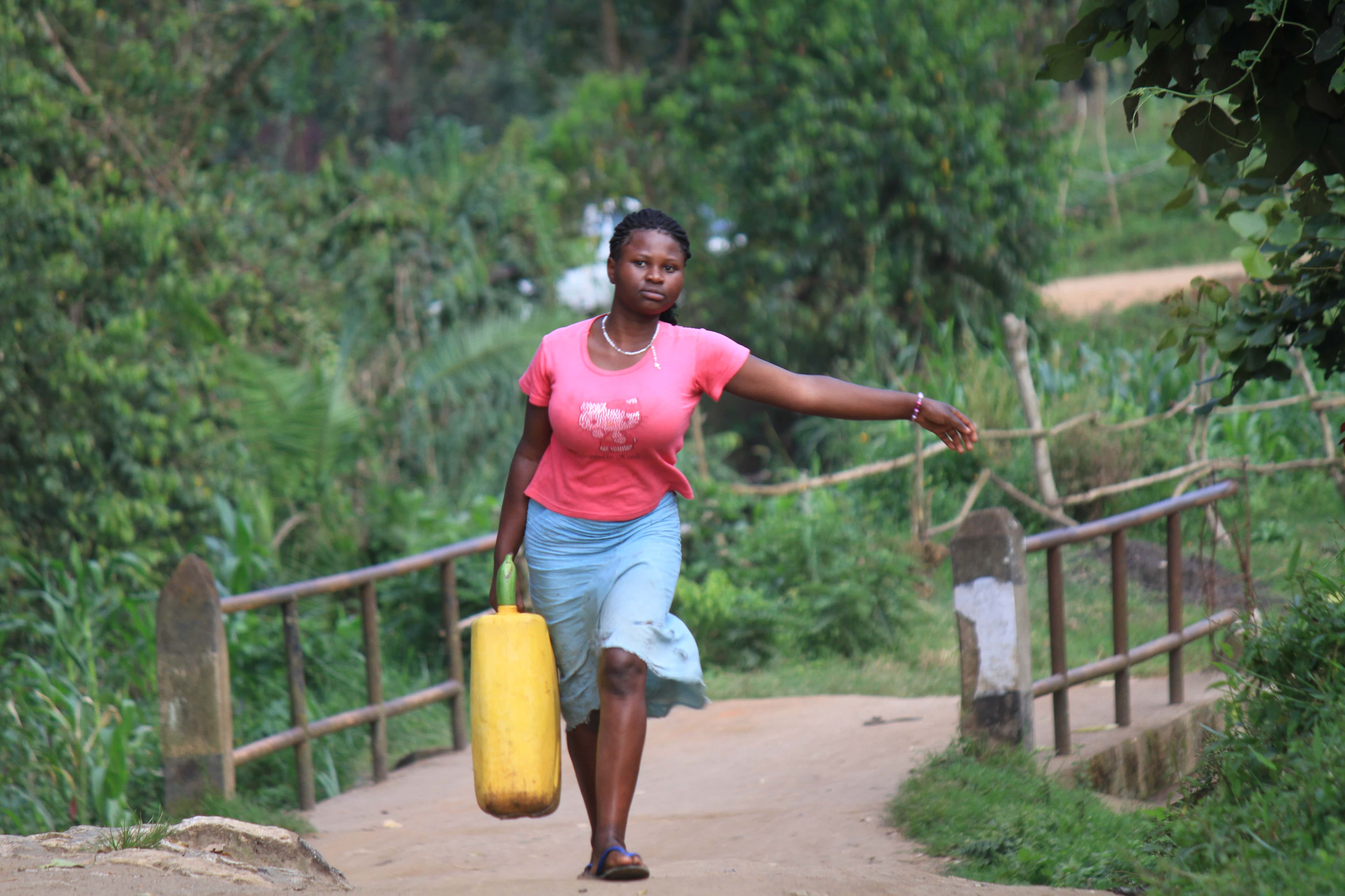 Woman carry jerrycan with water, wears pink shirt, blue dress