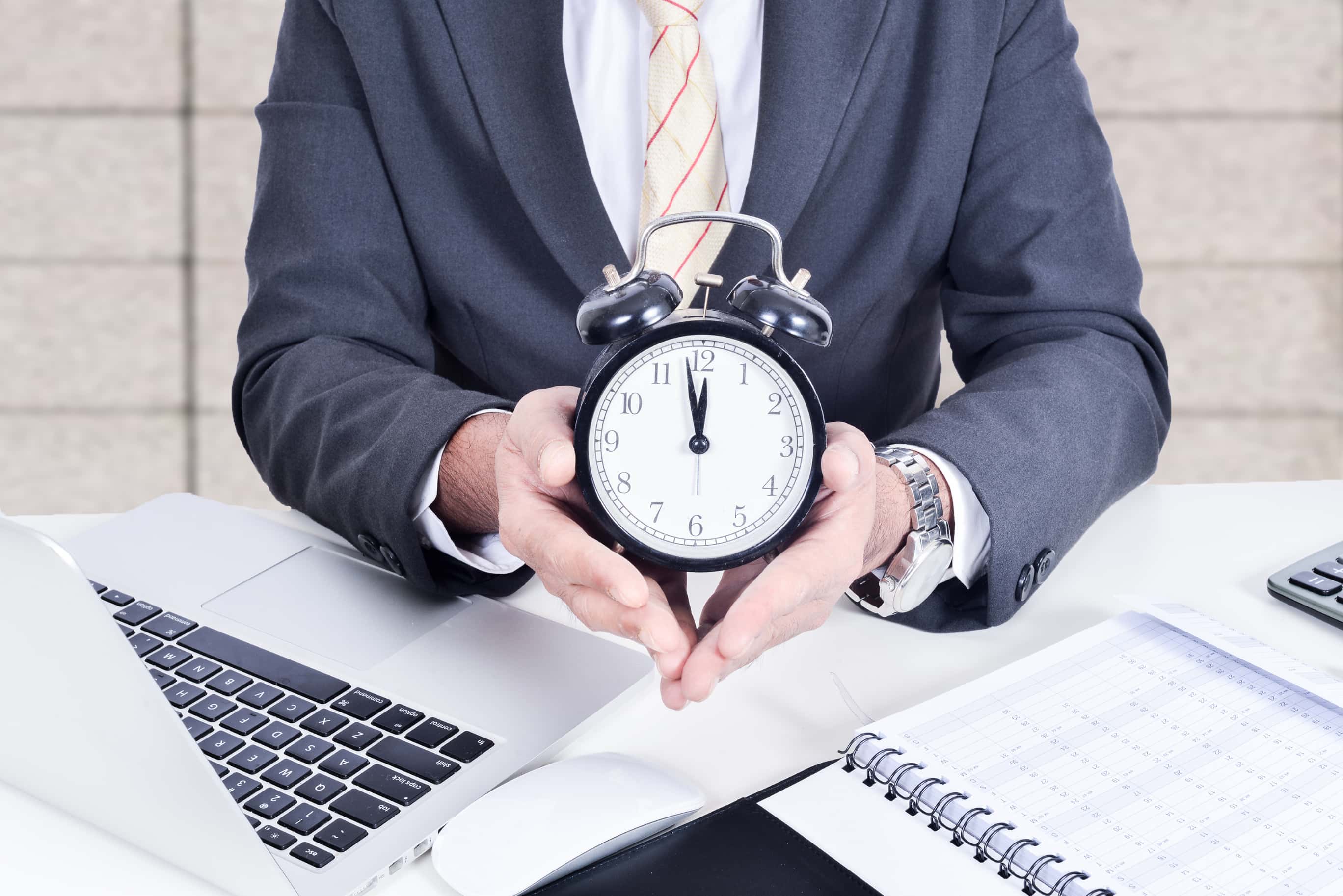 Midsection Of Businessman Holding Alarm Clock By Laptop On Desk