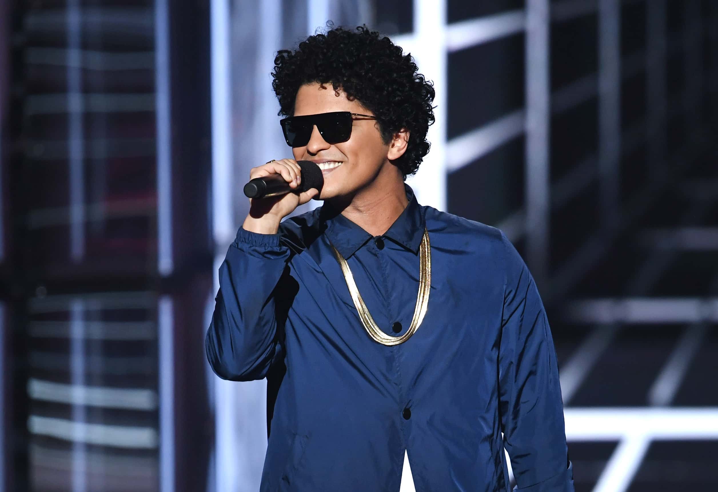 Bruno Mars speaks onstage during the Billboard Music Awards at MGM Grand Garden Arena in Las Vegas, Nevada. (Photo by Kevin Winter/Getty Images), wearing blue shirt and sunglasses
