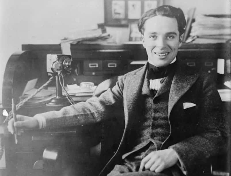 Grayscale Portrait Photo of the actor Charlie Chaplin, sitting on a chair holding a fountain pen, wearing dark suit, smiling