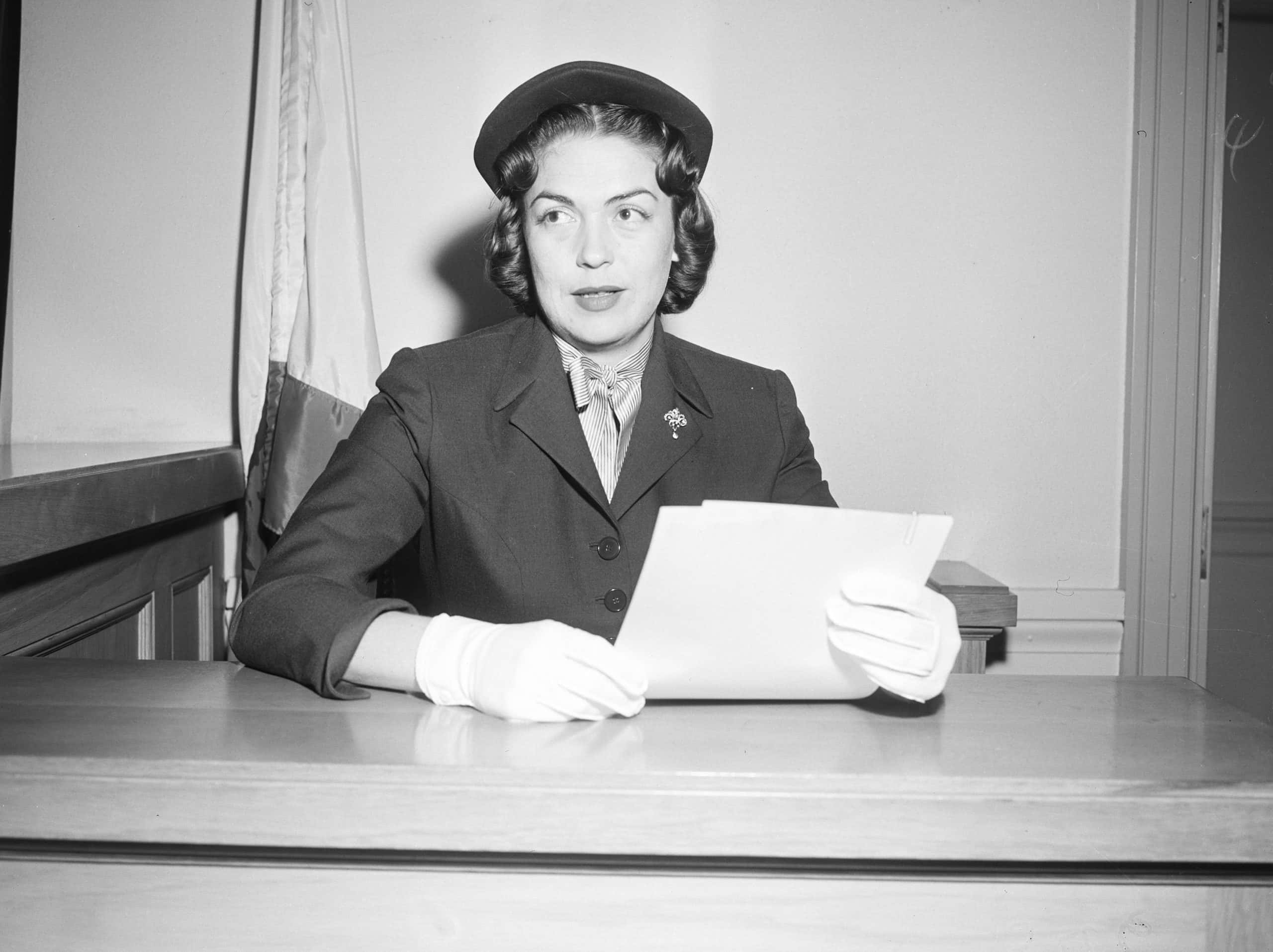 Esperanza Wayne sitting on a court table holding documents, wearing dark dress, dark hat