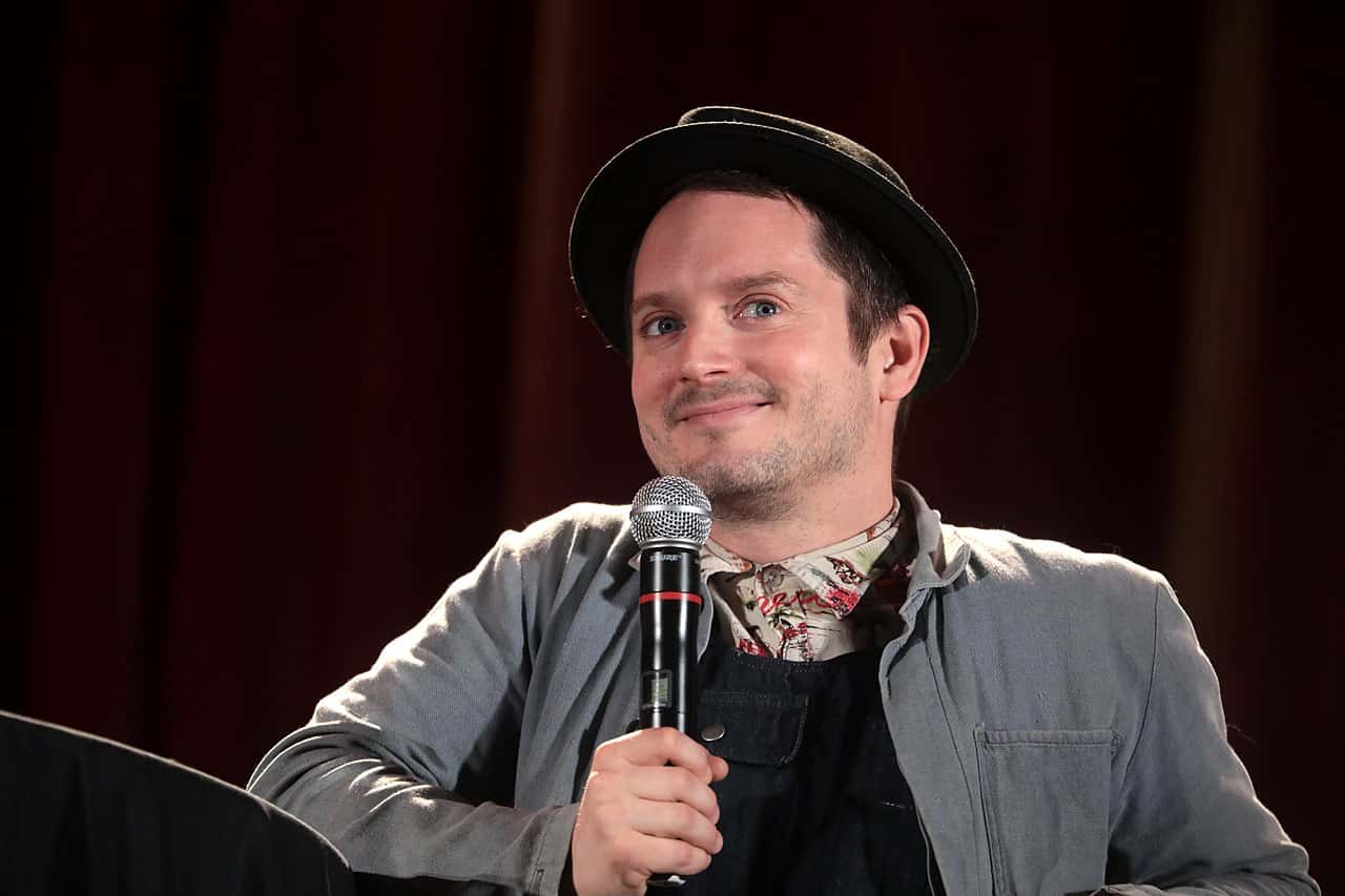 Elijah Wood wearing a grey jacket and a black hat