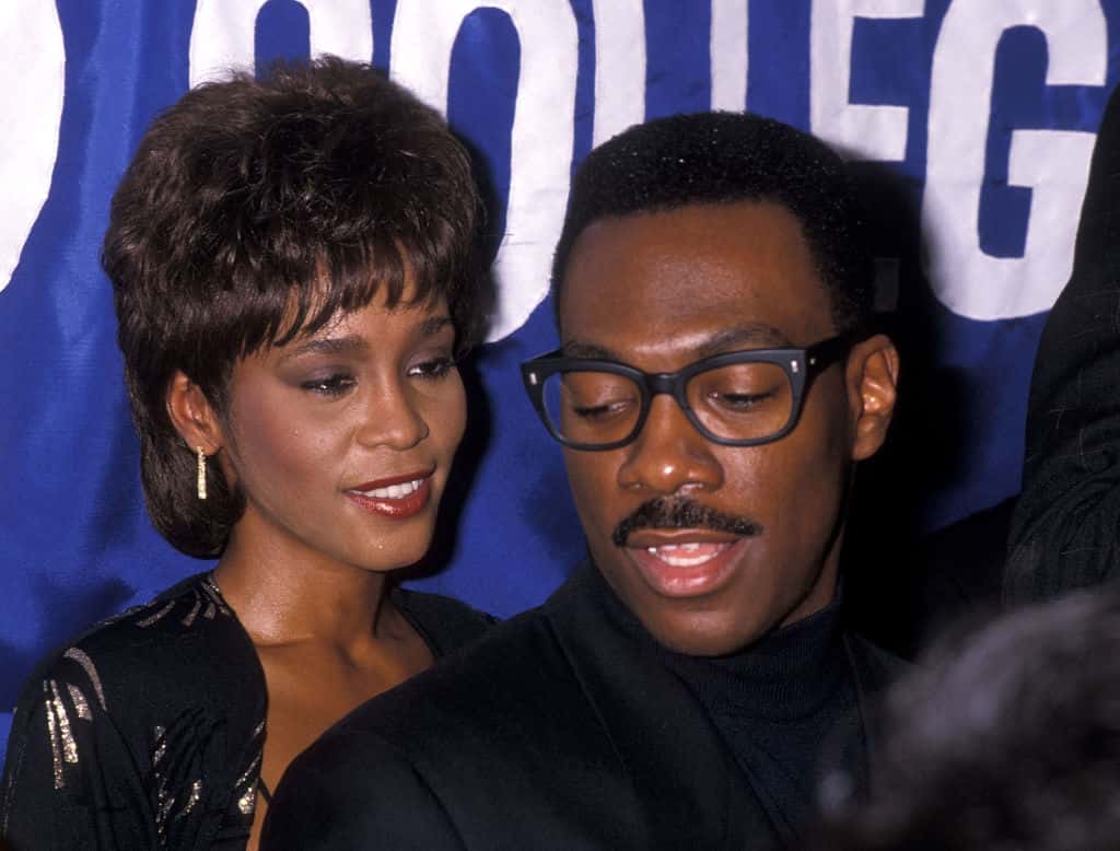 Whitney Houston in black dress and Eddie Murphy in black suit are talking and looking at side - 1989