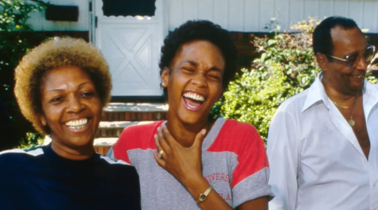 Screenshot: Whitney Houston wearing grey shirt is laughing with parents next to her  - from Whitney (2018)
