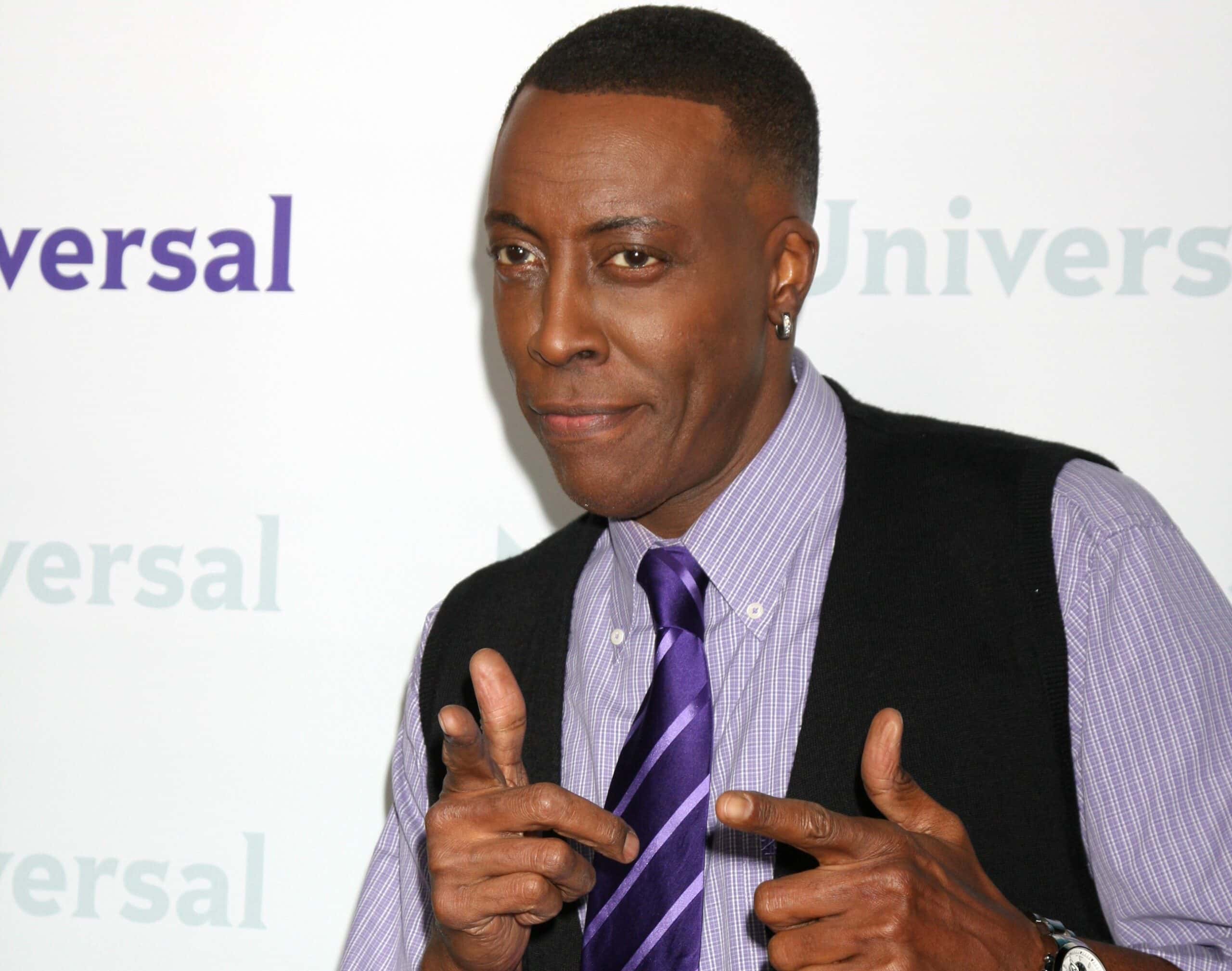 Arsenio Hall wearing purple shirt and tie is pointing and smiling at side - 2012