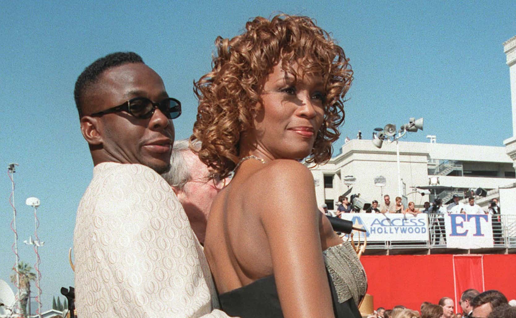 Whitney Houston wearing black dress and sunglasses and Bobby Brown wearing white jacket are looking and smiling outside - 1998