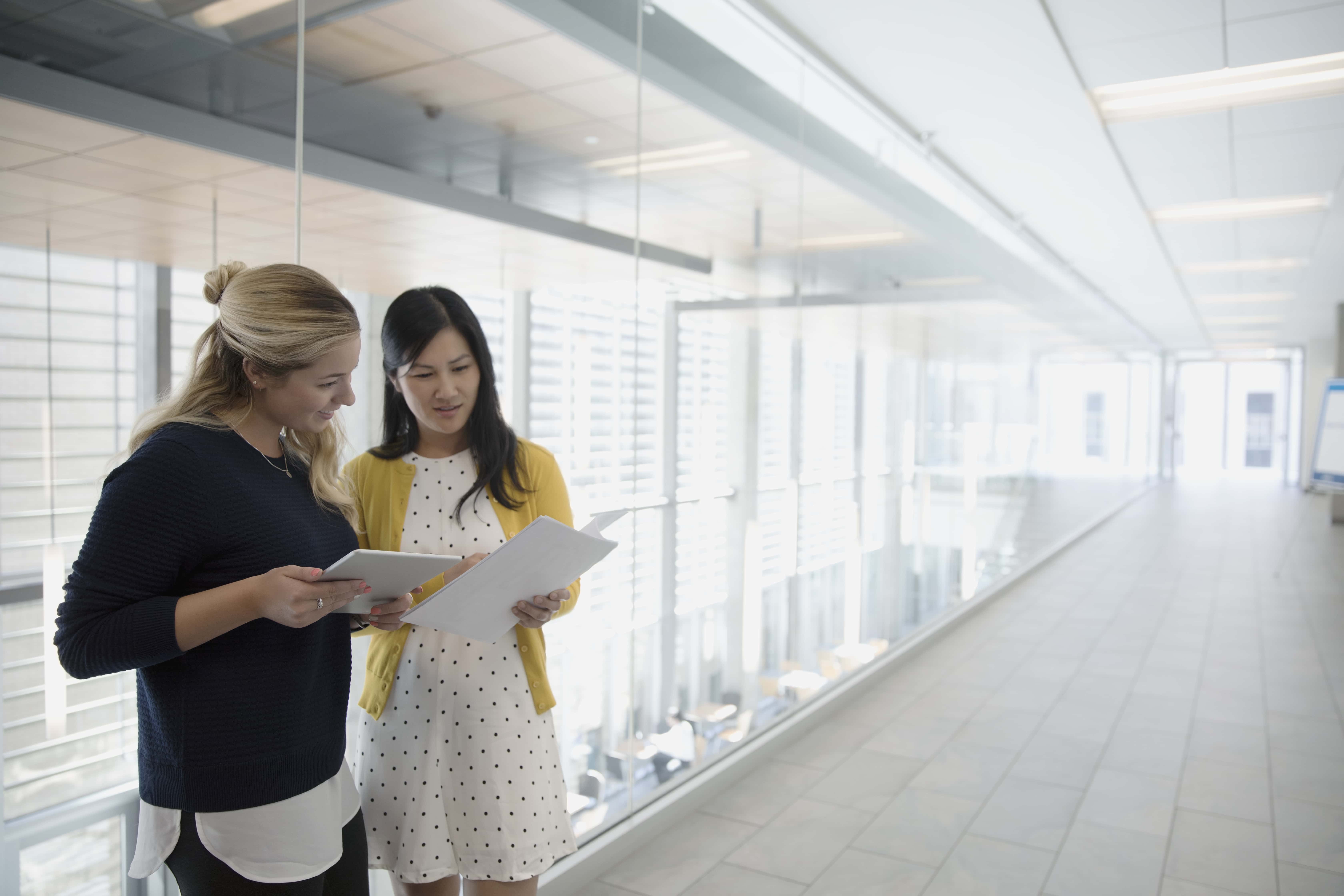 Businesswomen using digital tablet, discussing paperwork in office corridor.