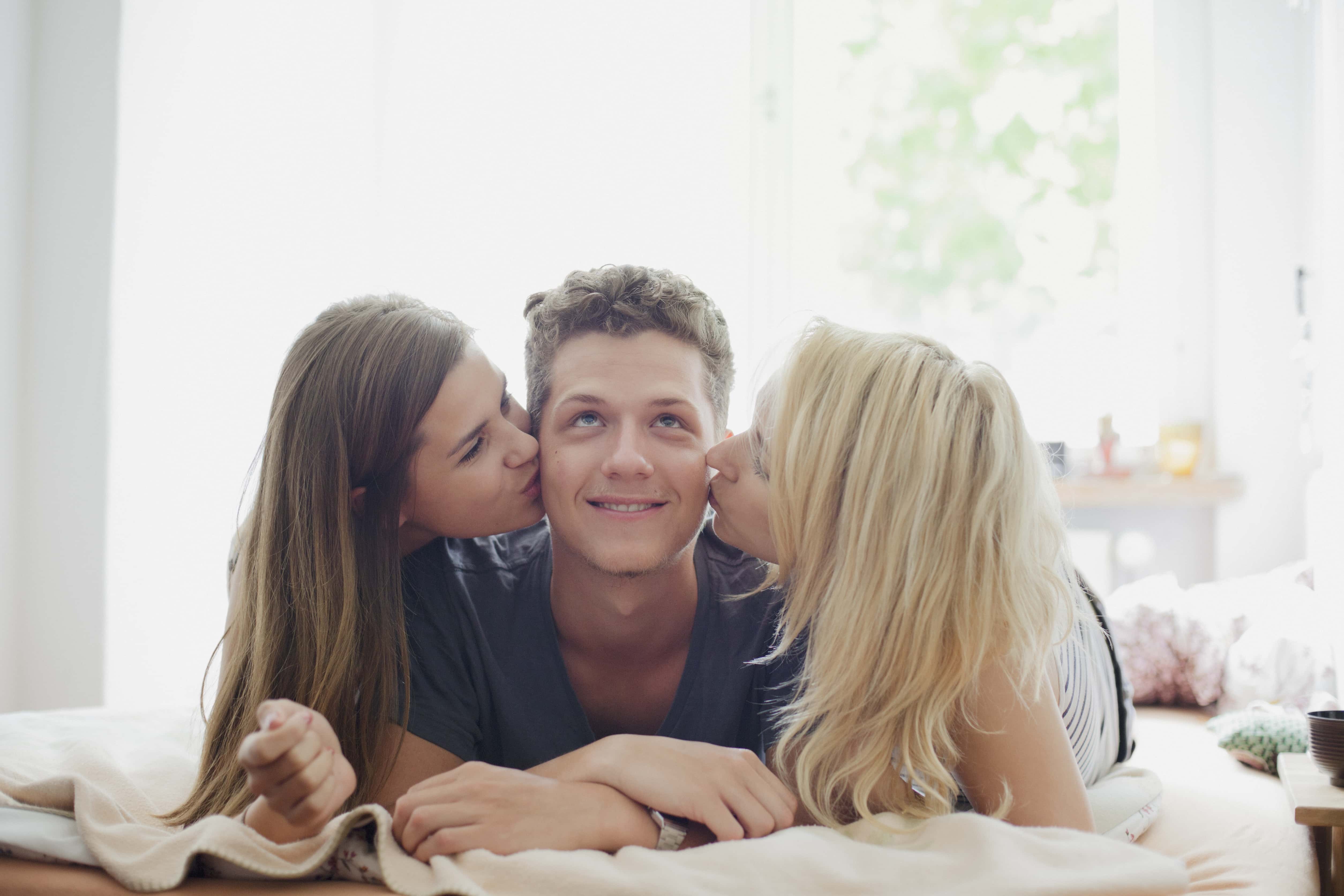 Young female friends kissing handsome man lying on bed at home.