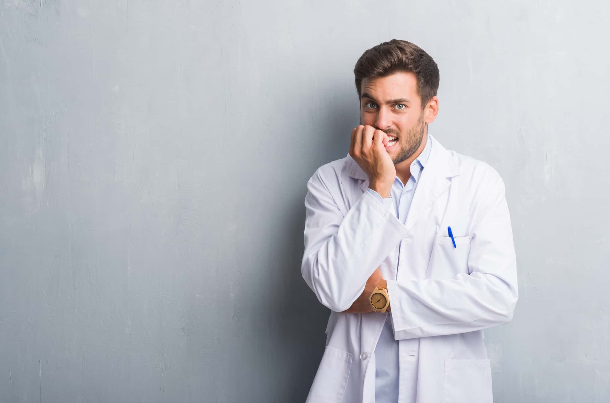 Handsome man wearing white coat looking stressed against grey background.