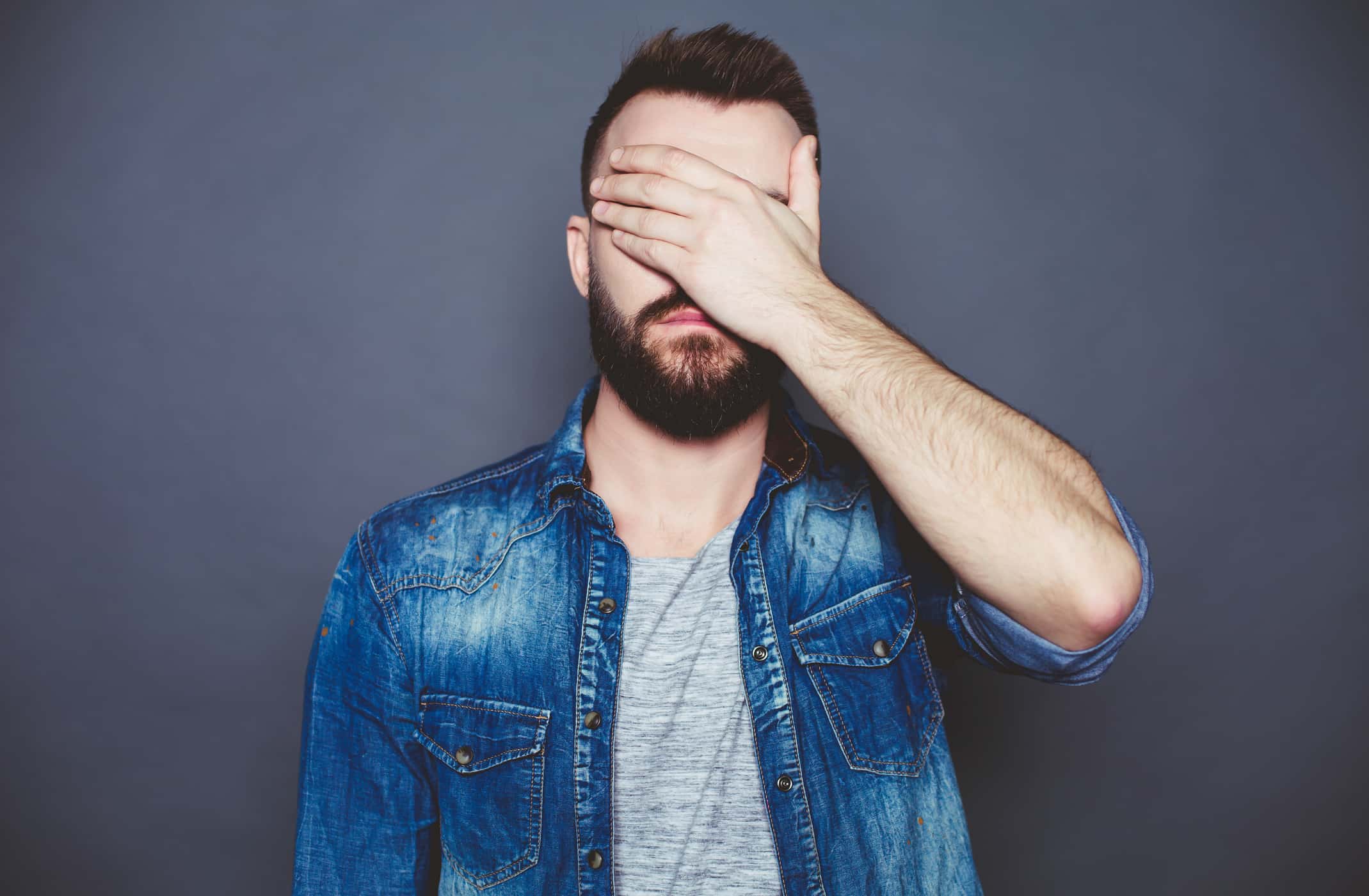 A young man in a denim shirt closing his eyes with his hand.