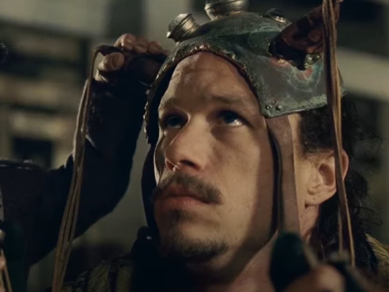 Screenshot: Heath Ledger with a long hair wearing grey jacket and helmet 
