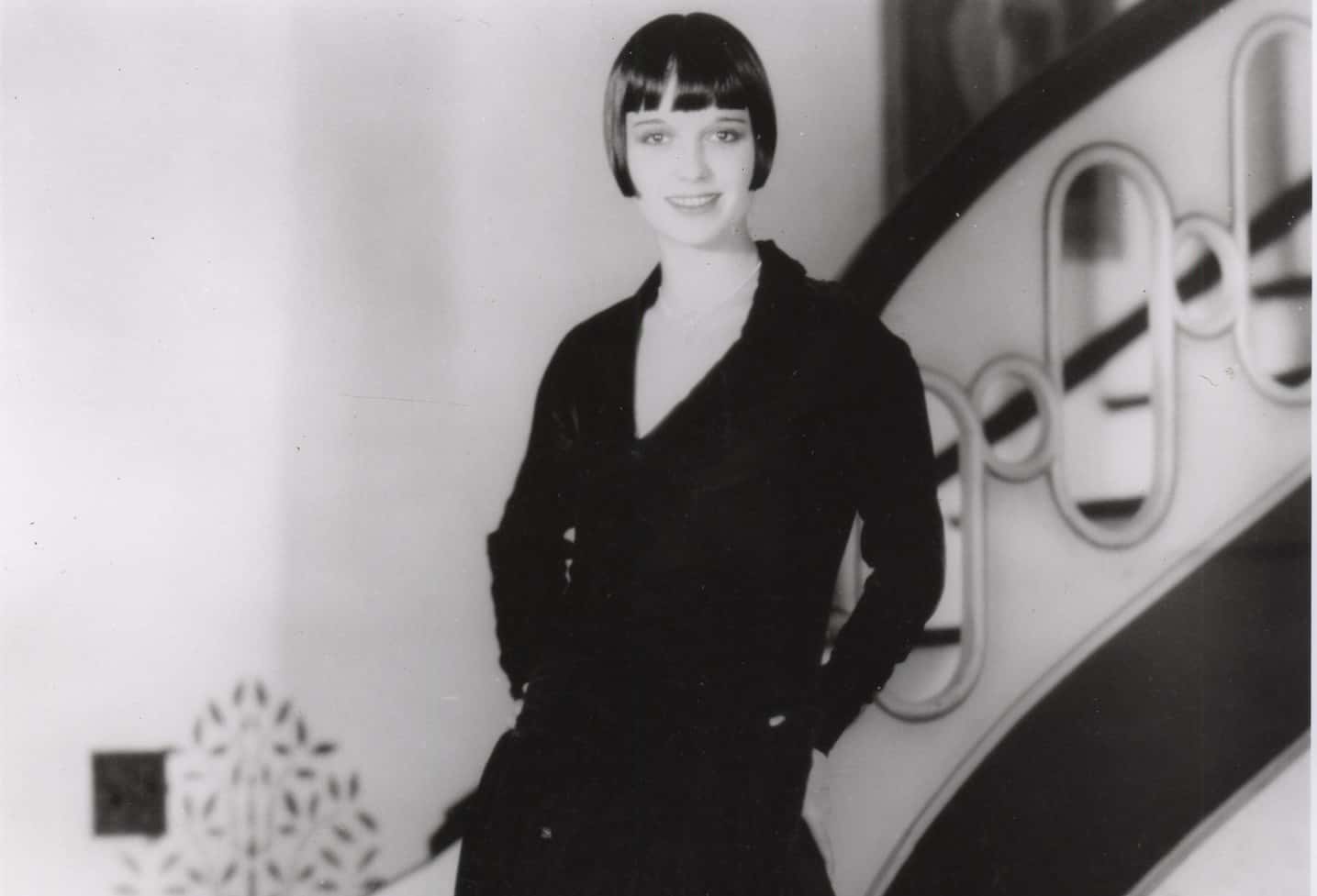 Grayscale Portrait Photo of the actress Louise Brooks leaning on stairs, wearing a black dress, smiling