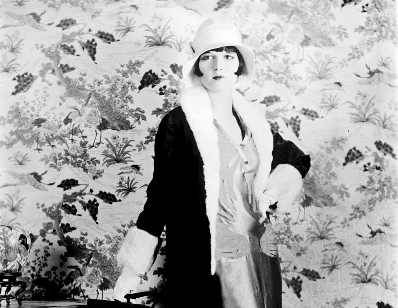 Grayscale Portrait Photo of the actress Louise Brooks, wearing a black coat, light colored dress and a white hat