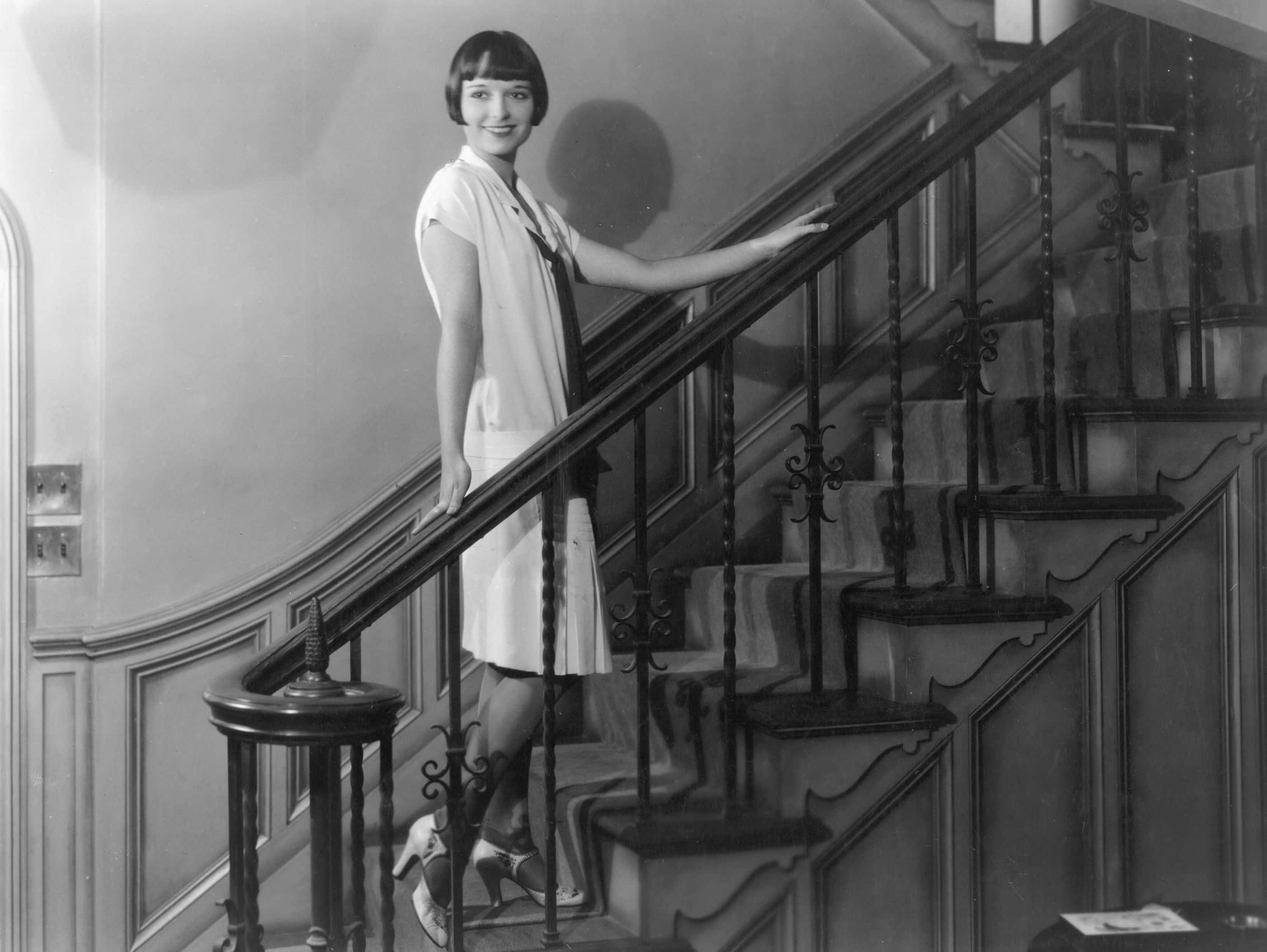  Grayscale Photo of Actress Louise Brooks, standing on stairs, wearing white dress, smiling