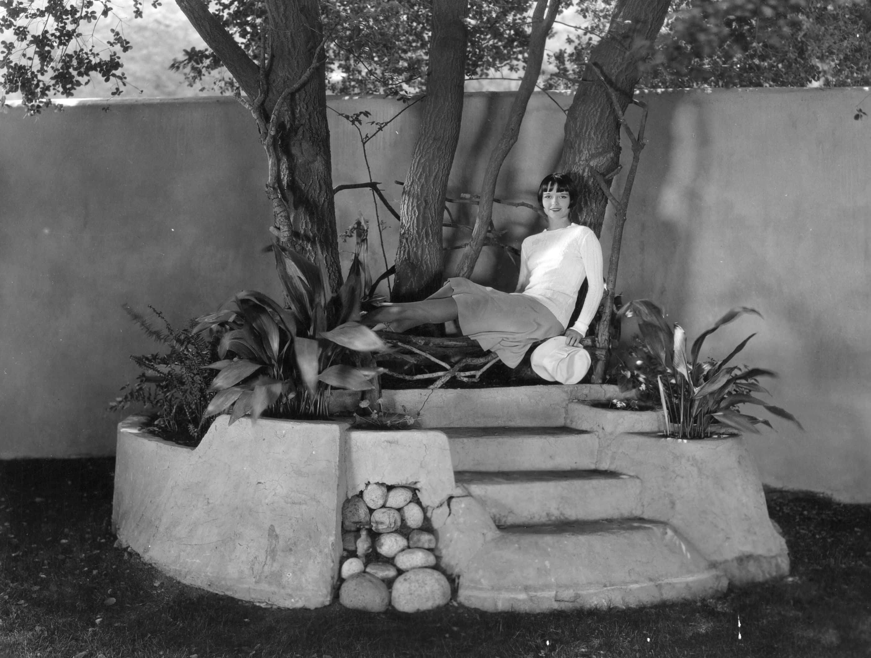 Actress Louise Brooks, sitting relaxes in her garden, wearing light colored outfit, smiling