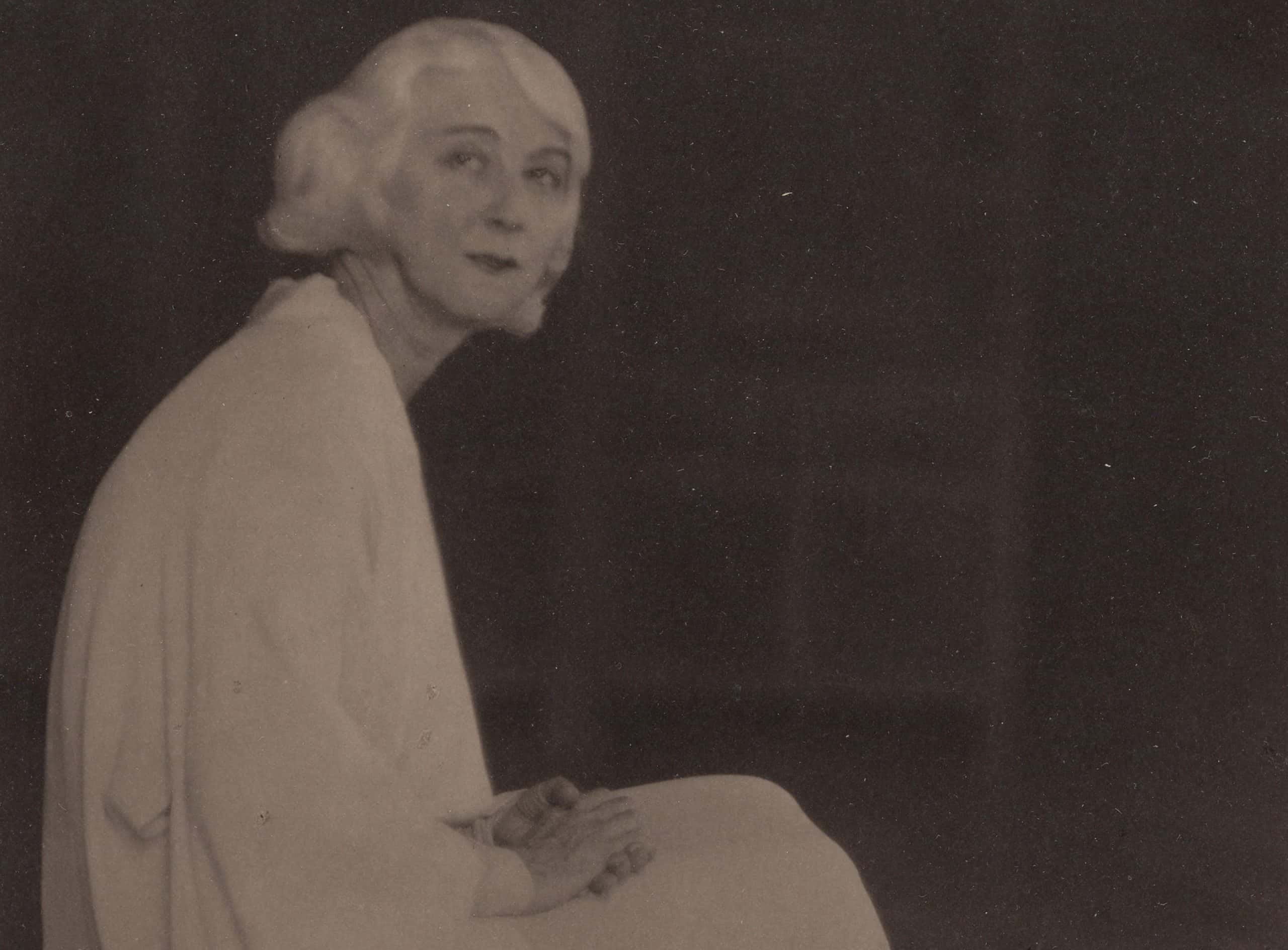 Ruth St. Denis sitting on a chair, wearing a white dress, facing the camera