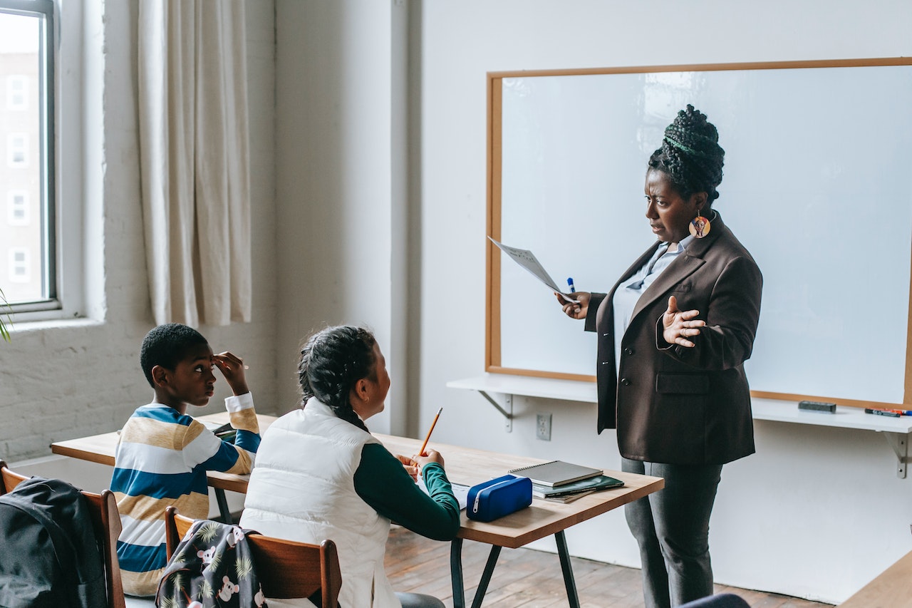 Black teacher talking to the kids seating in the classroom and holding a paper.