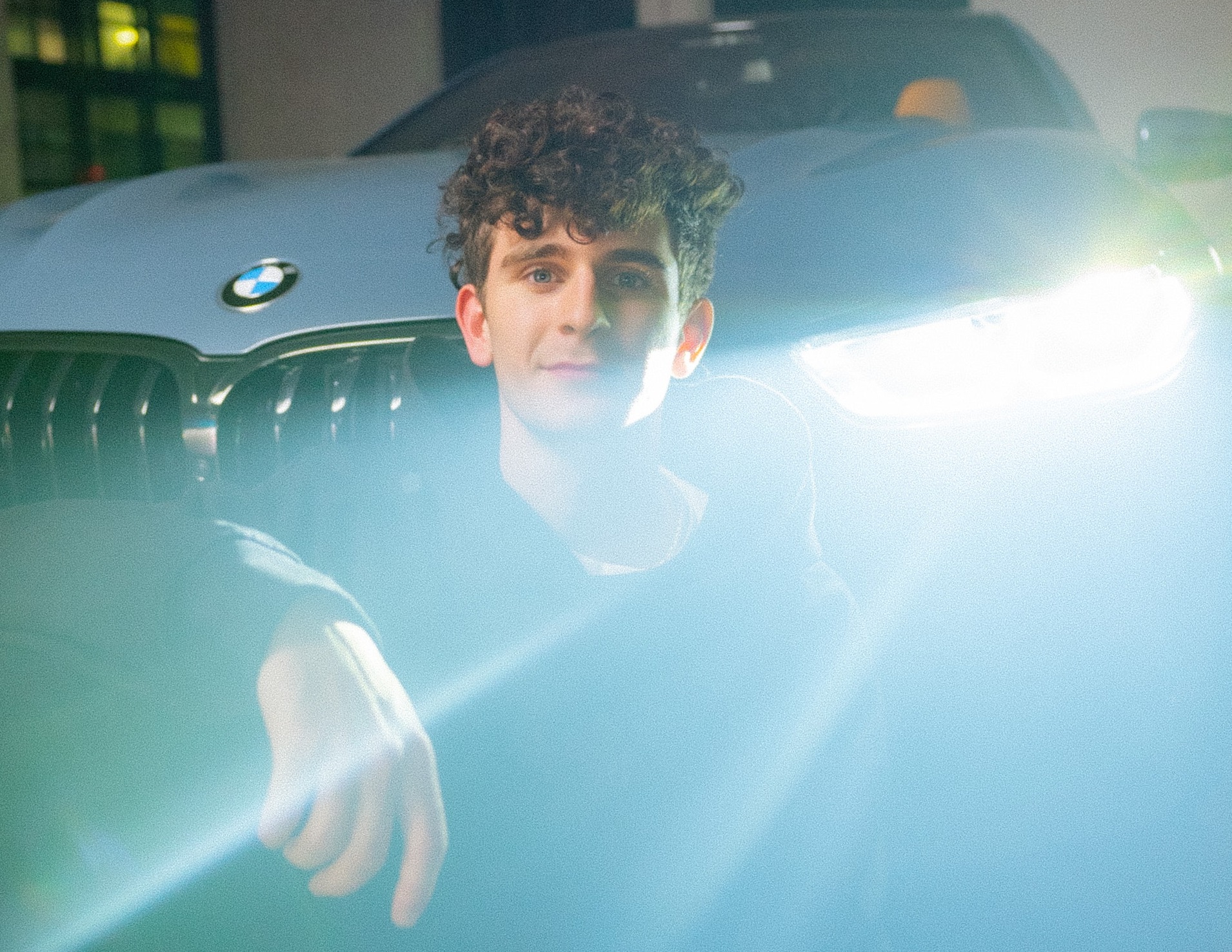 A man sitting in front of a BMW car with headlight on.
