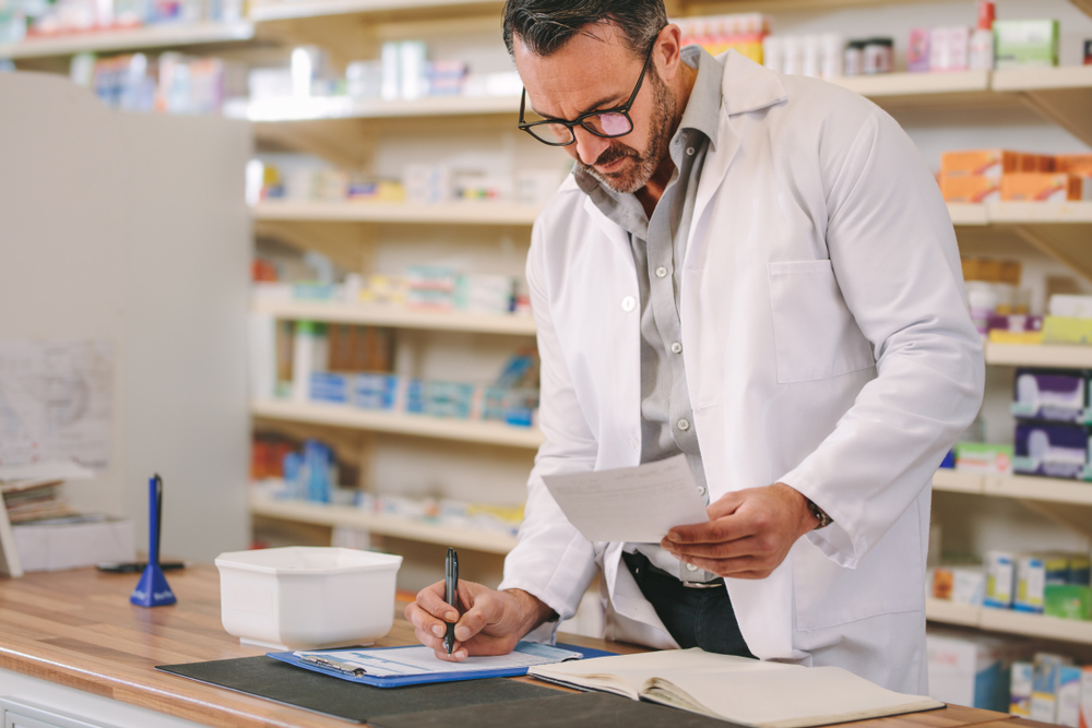 Male pharmacist writing prescription pharmacy counter in white uniform
