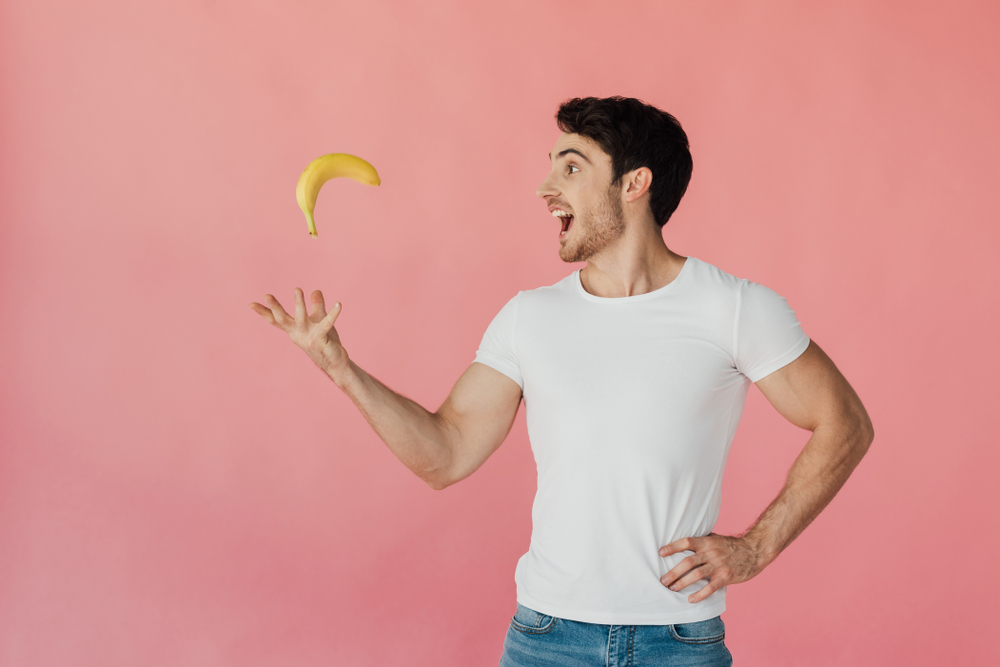 Smiling muscular man in white t-shirt throwing up banana 