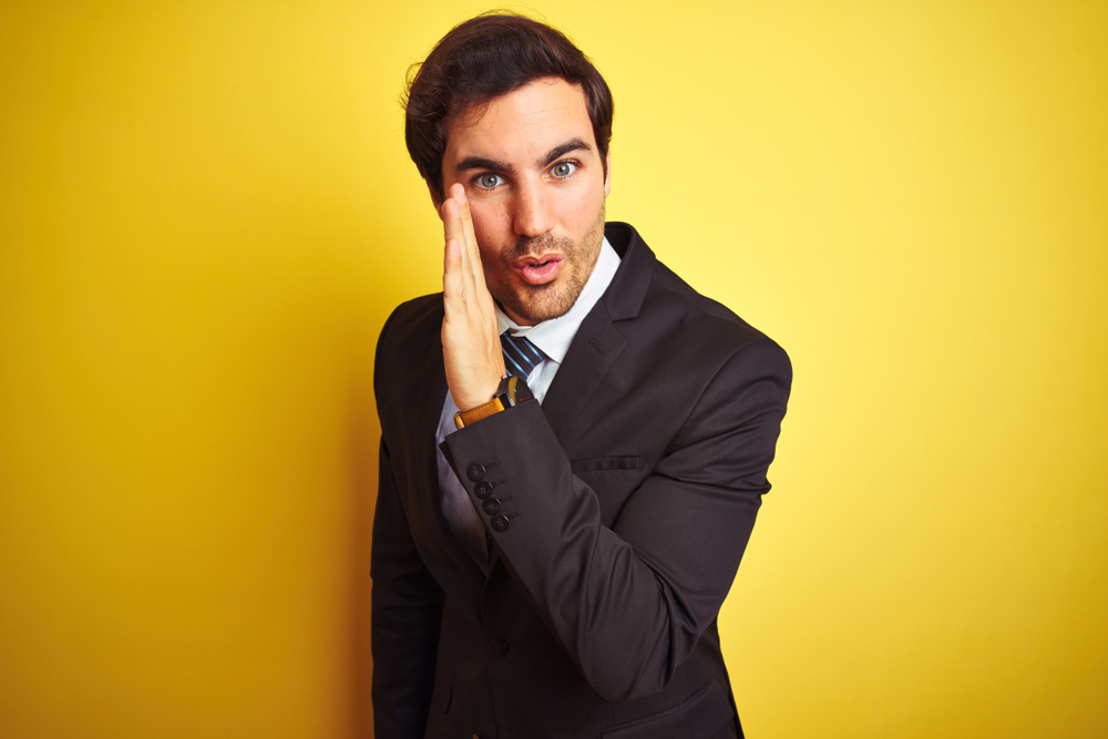 Businessman standing over yellow background with hand on mouth telling secret
