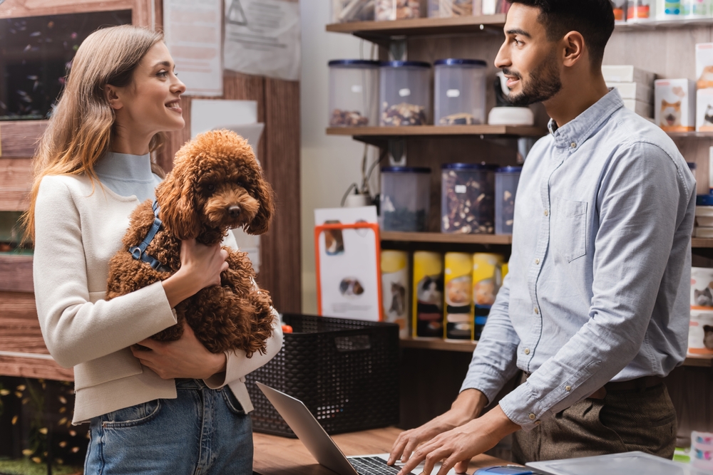 Side view of smiling woman holding poodle near Arabian seller in pet shop