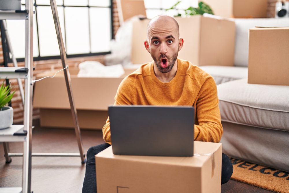 Young bald man with beard moving to a new house using laptop scared and amazed with open mouth for surprise, disbelief face