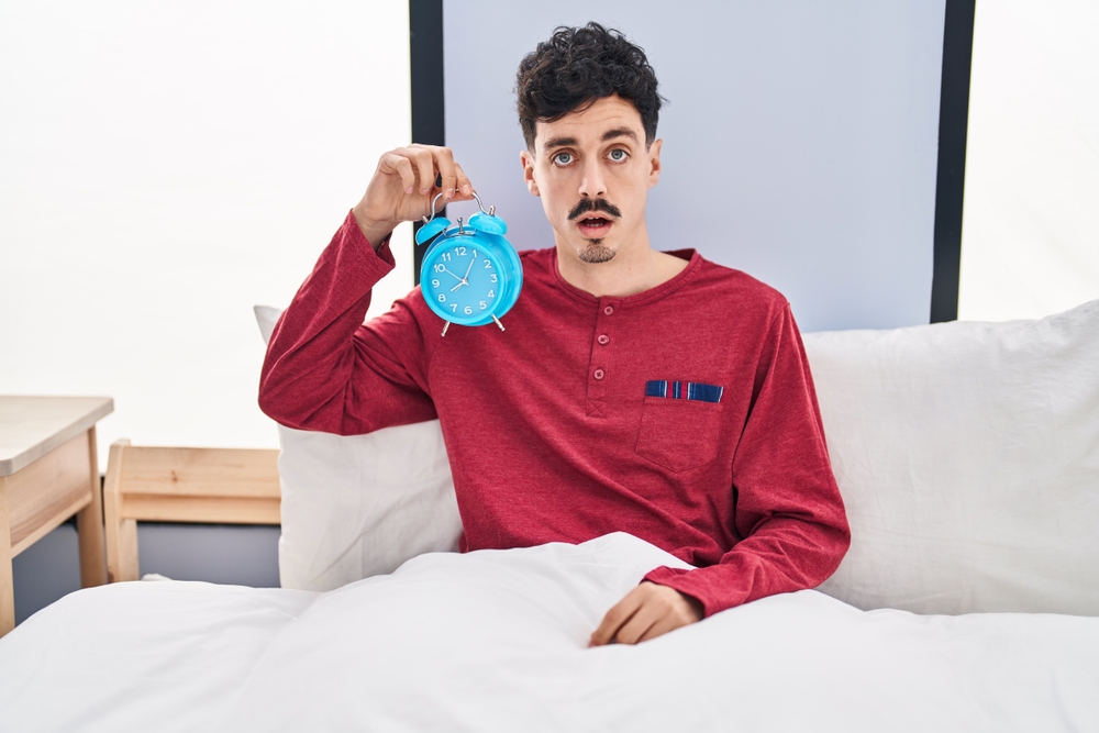 Shocked man just woke up in bed holding blue alarm clock