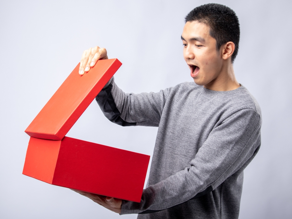 A portrait of an Asian man wearing a gray sweater showing a surprised expression while opening a red gift box