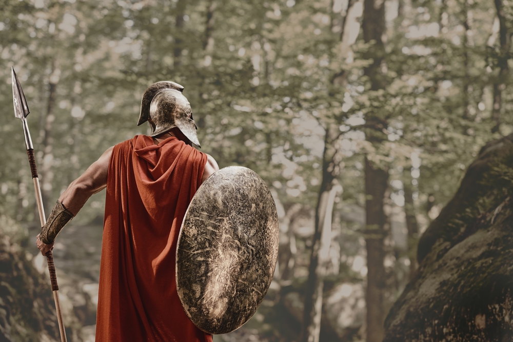Back view of man in Spartan helmet and red long cloak standing with round shield and spear in hands looking away