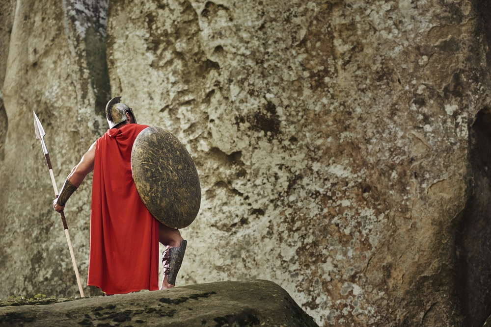 Back view of victorious warrior in long red cloak and gladiator helmet posing with heavy shield