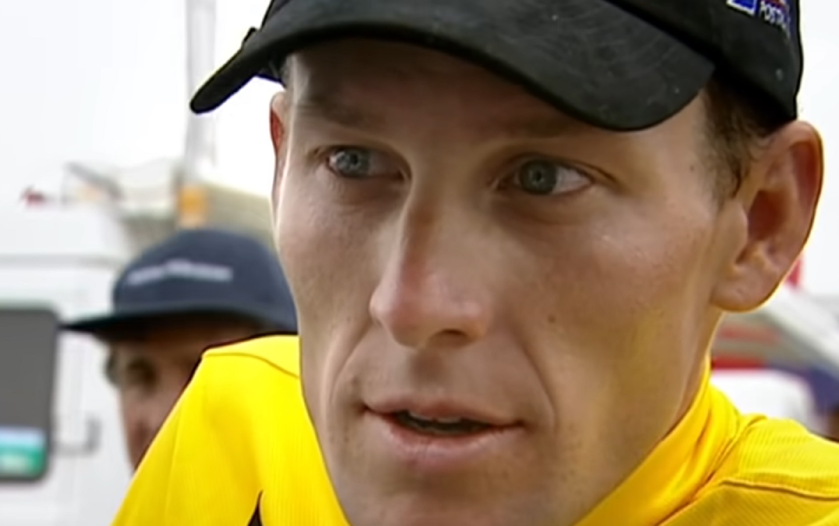 Screenshot: Lance Armstrong wearing yellow shirt and black hat is looking at side -from The Armstrong Lie (2013)