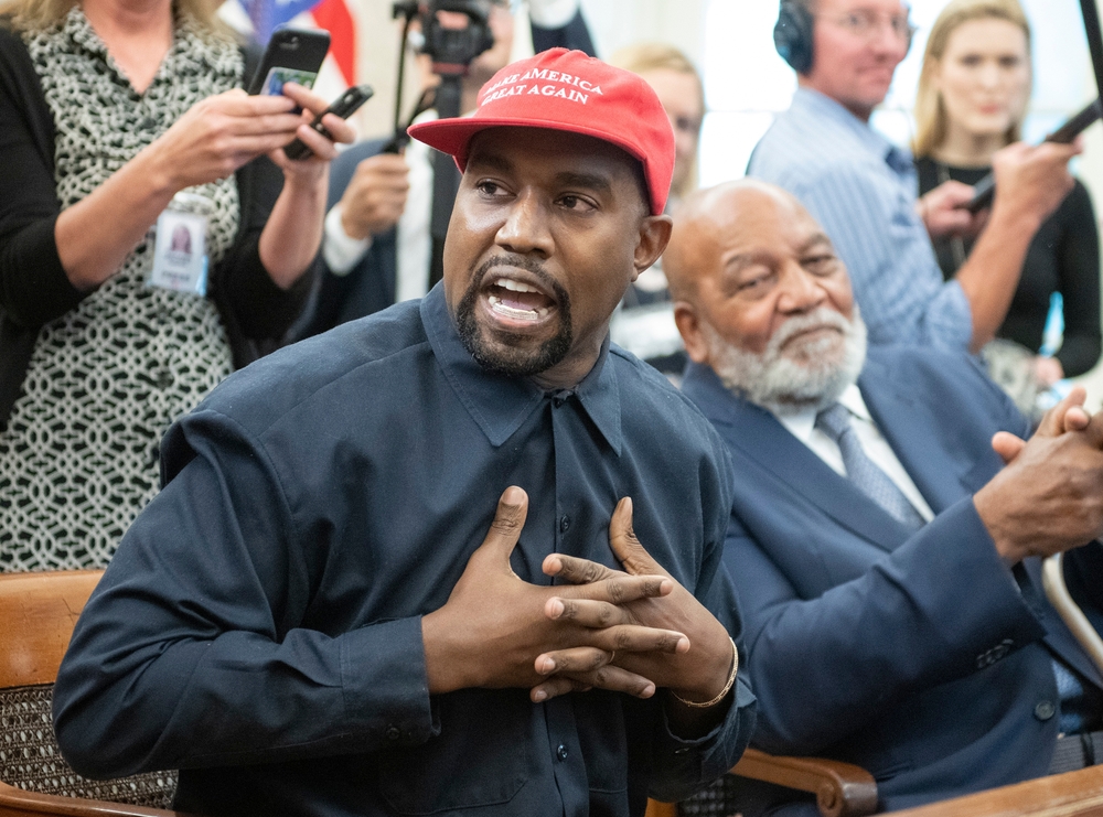 Kanye West speaks with his hands crossed placed at the chest with black shirt and red hat